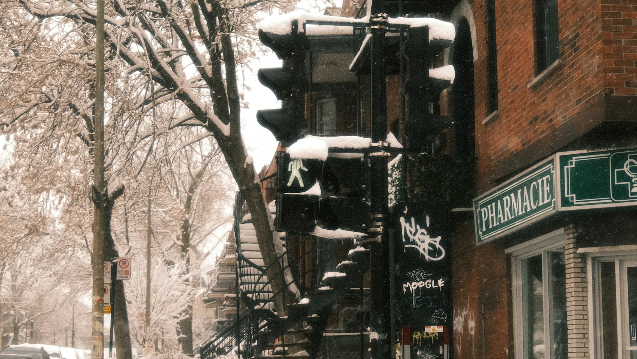 Coin de rue enneigé à Montréal, avec une pharmacie et un escalier extérieur en colimaçon.